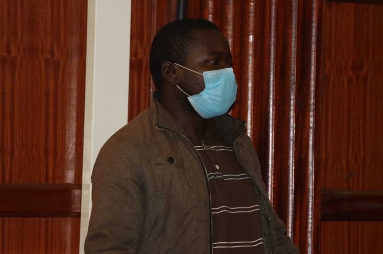 Le Kenyan Augustine Wanyonyi lors de son procès en décembre 2021. Il se tient très droit. Il est habillé en brun et porte un masque anti-covid. On ne voit que son buste. Derrière lui, des boiseries horizontales.