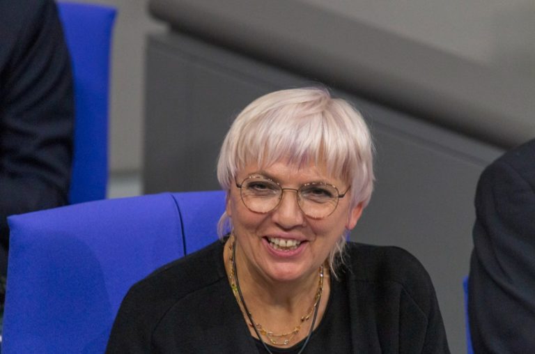 Photo de Claudia Roth, environ 65 ans, est assise, et lève les yeux vers l'objectif en souriant. Elle a les cheveux courts platine, et porte des lunettes.