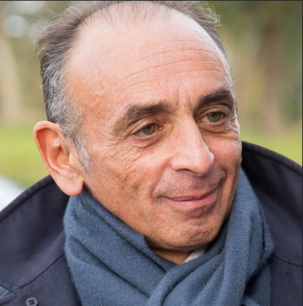 Eric Zemmour, souriant, a la tête tournée vers la droite. Il a environ 60 ans, et est un peu dégarni. On voit qu'il porte une écharpe.