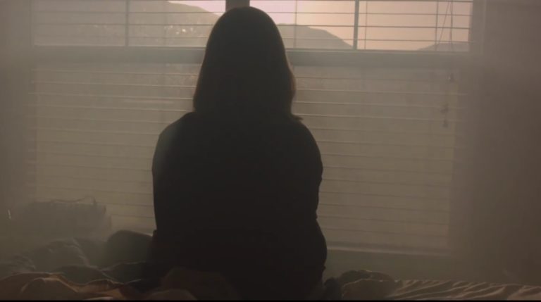 Photo extraite du fil Light your world. Une jeune femme est assise de dos dans la pénombre. Face à elle, une grande et douce lumière passe à travers une fenêtre.