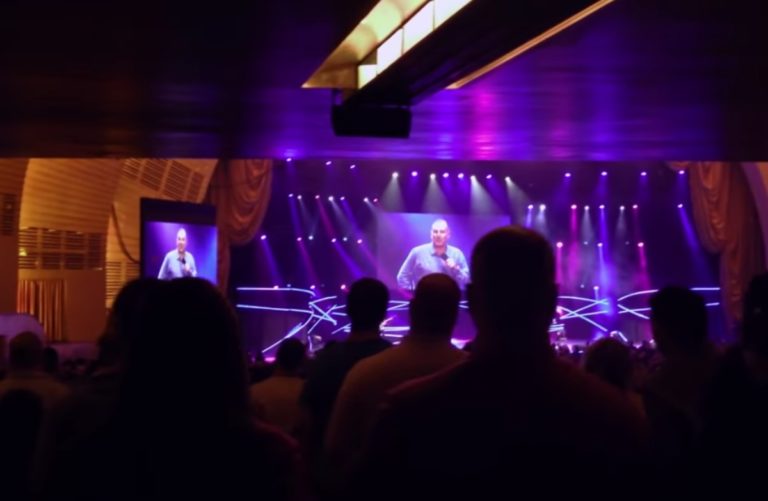 Dans une vaste salle, une foule regarde vers un écran géant situé au-dessus d'une scène. On y voit le psteur Brian Houston.