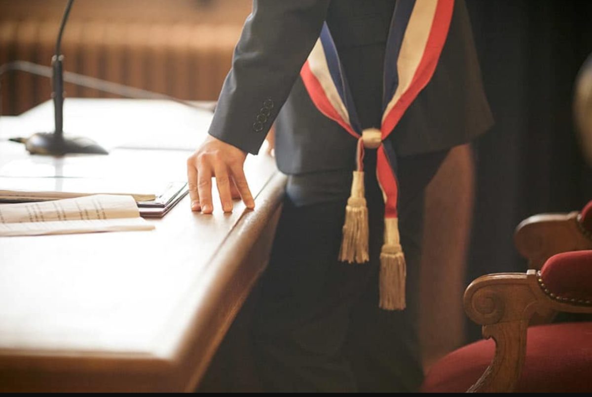 La main d'un homme portant l'écharpe tricolore est osée sur un vaste bureau.