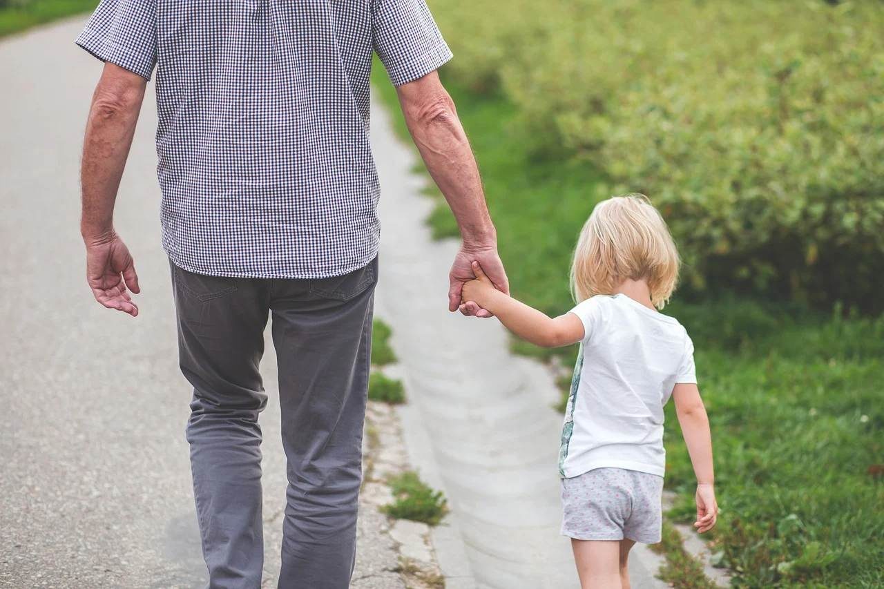 De dos, un homme tient sa petite fille blonde par la main. Ils sont au bord d'une route de campagne. L'enfant semble avoir environ 5 ans.