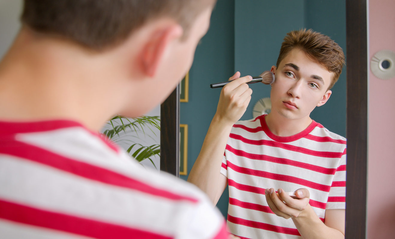 Un adolescent androgyne se maquille en se regardant dans un miroir.