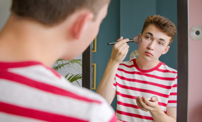 Un adolescent androgyne se maquille en se regardant dans un miroir.