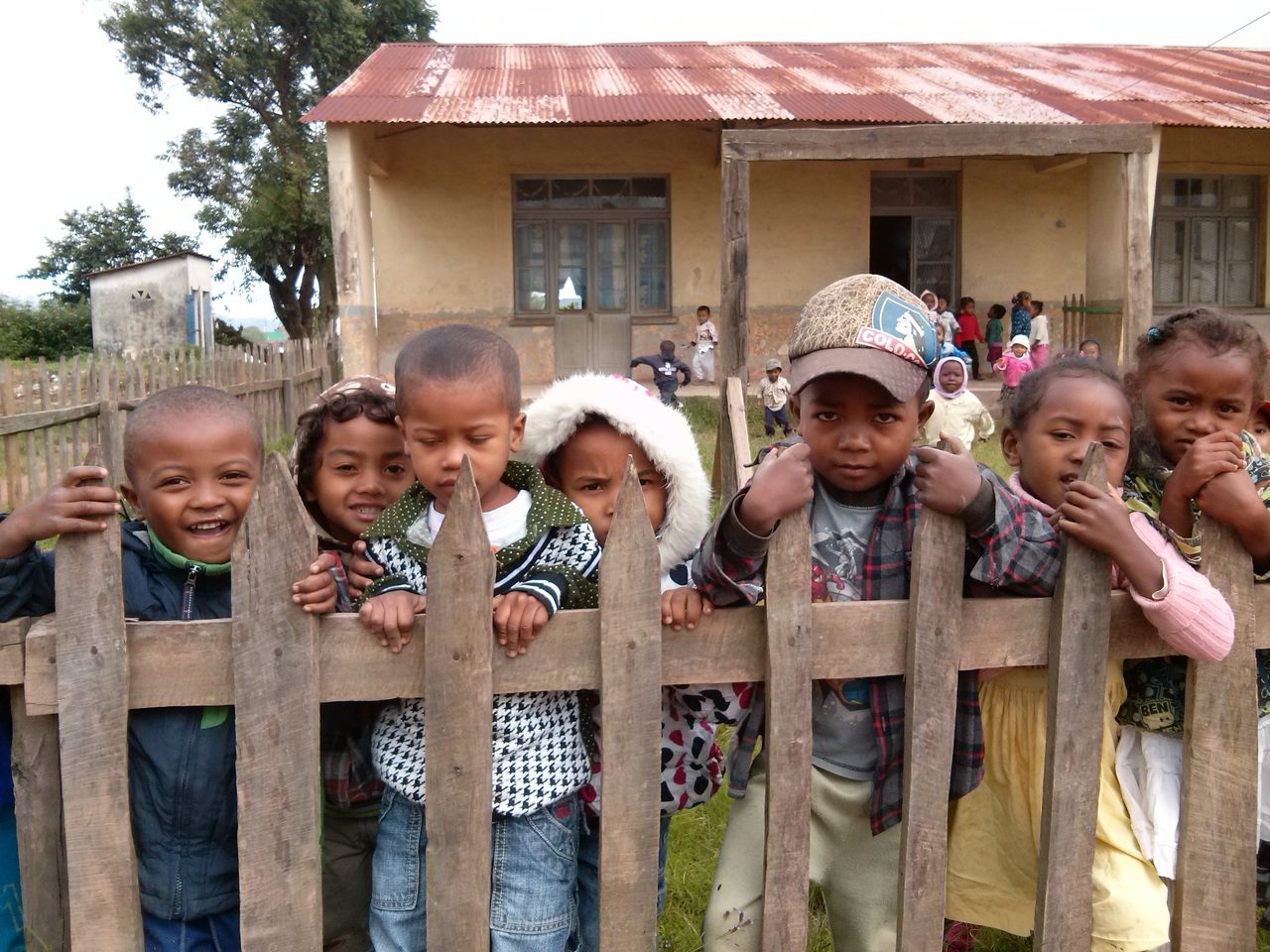 Des enfants se pressent derrière une clôture en bois. Derrière eux se trouve un petit bâtiment insalubre avec un toit en tôles. D'autres enfants sont en train de jouer sur le perron.