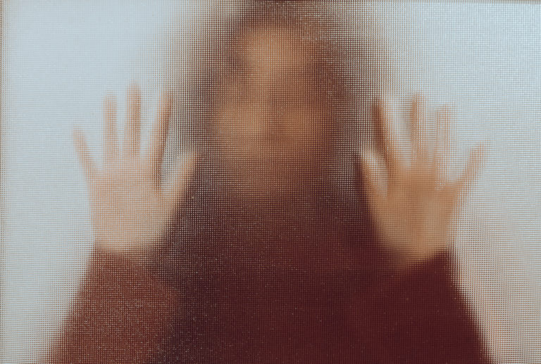 Une femme pose ses mains sur une vitre brouillée. Elle est derrière et elle est difficile à distinguer. L'image renvoie une forte impression d'enfermement et de censure.