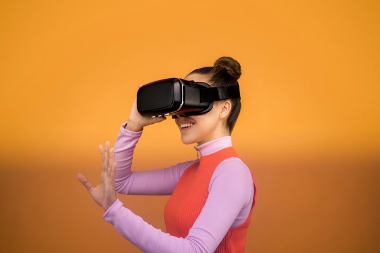 Une femme porte un casque de réalité virtuelle. Elle sourit et semble essayer de toucher quelque chose près d'elle, qu'elle voir avec l'appareil.