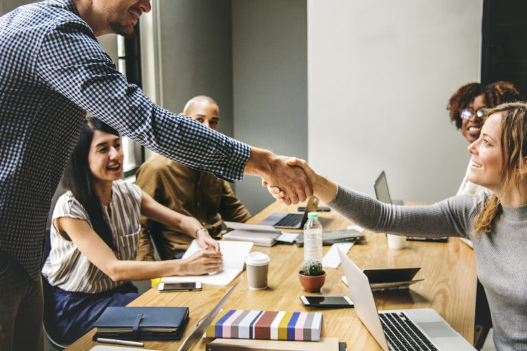 Plusieurs personnes sont assises autour d'une table où elles travaillent avec leurs ordinateurs, de toute évidence dans une entreprise. Au premier plan, un homme, debout, serre la main d'une femme qui est assise.