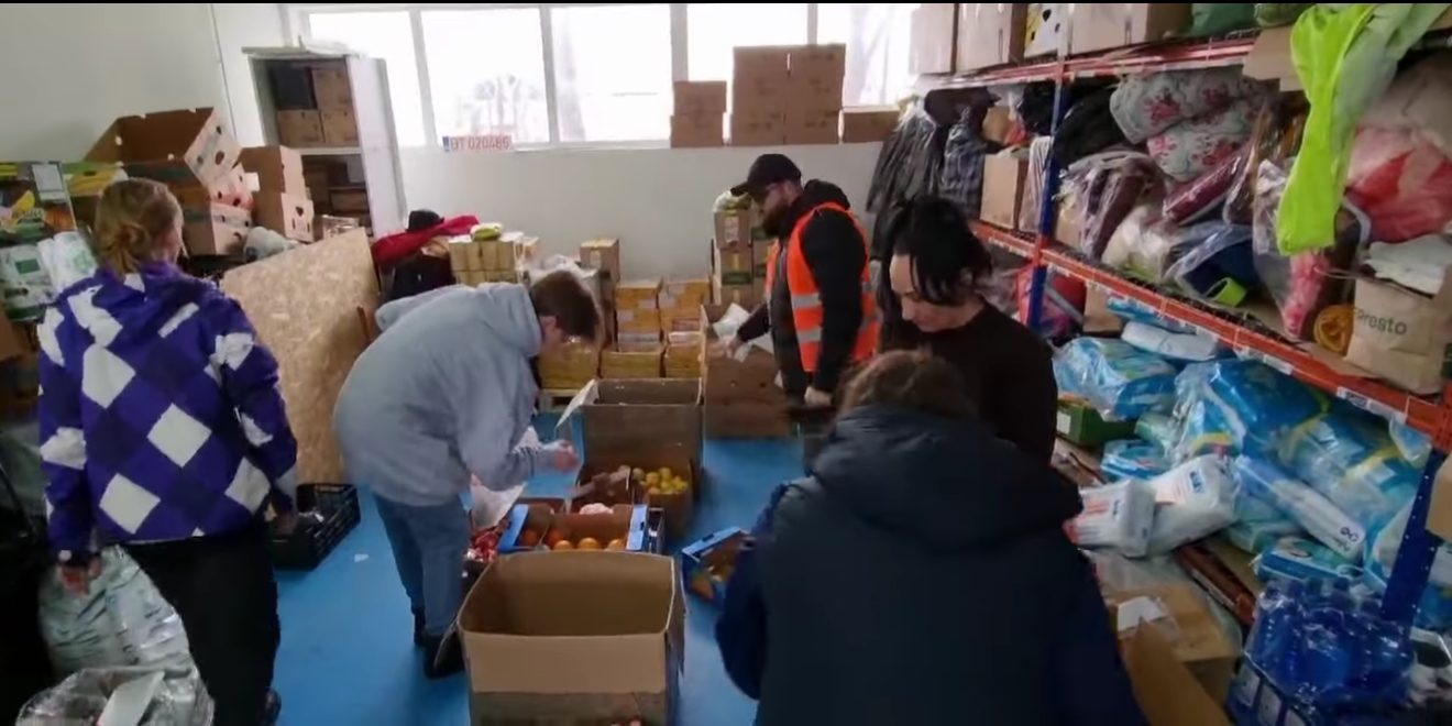 Un groupe de persnne s'activent dans un petits entrepôt à provision.s. Ils sont en habits d'hiver et l'entrepôt est visiblement une construction précaire.