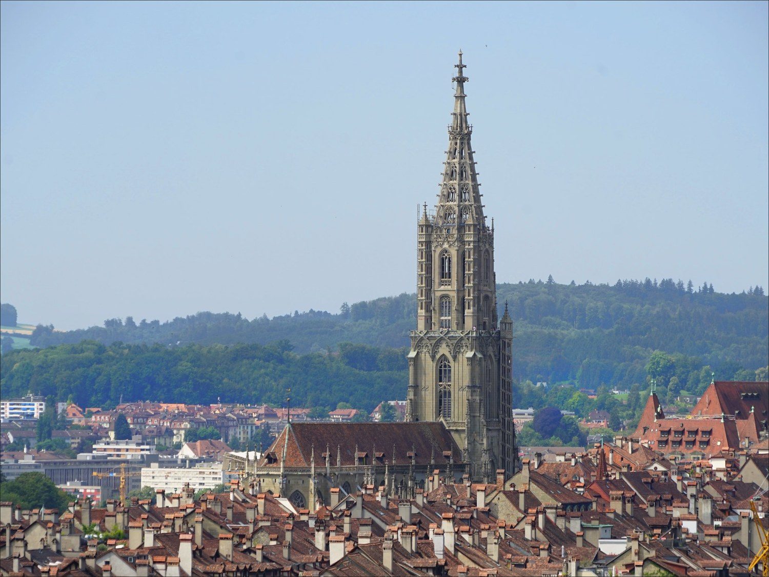 Vue de la cathédrale de Berne, avec une multitude de toits de maisons au premier plan. Sa flèche est très haute par rapport au reste du bâtiment, qui est moins massif.