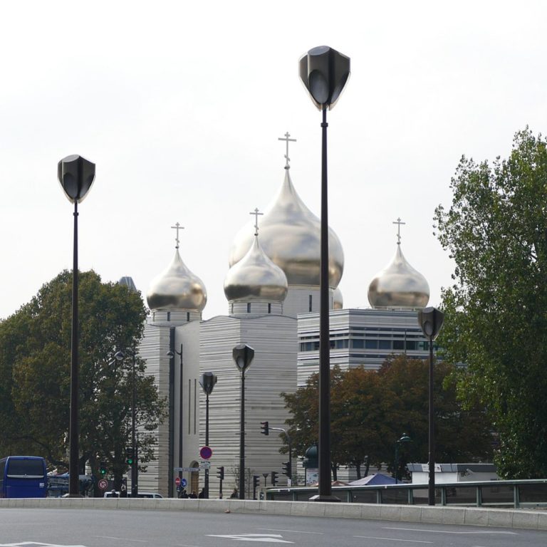 La cathédrale de la Sainte trinité à Paris, vue depuis une route. Elle est constituée d'un énorme bloc gris et surmontée de domes argentés.