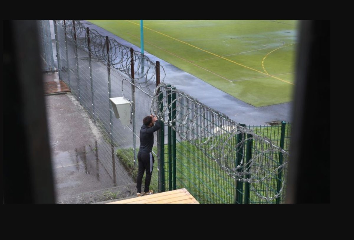 Un détenu regarde à travers un grillage d'une cour de prison, les deux coudes appuyés dessus.