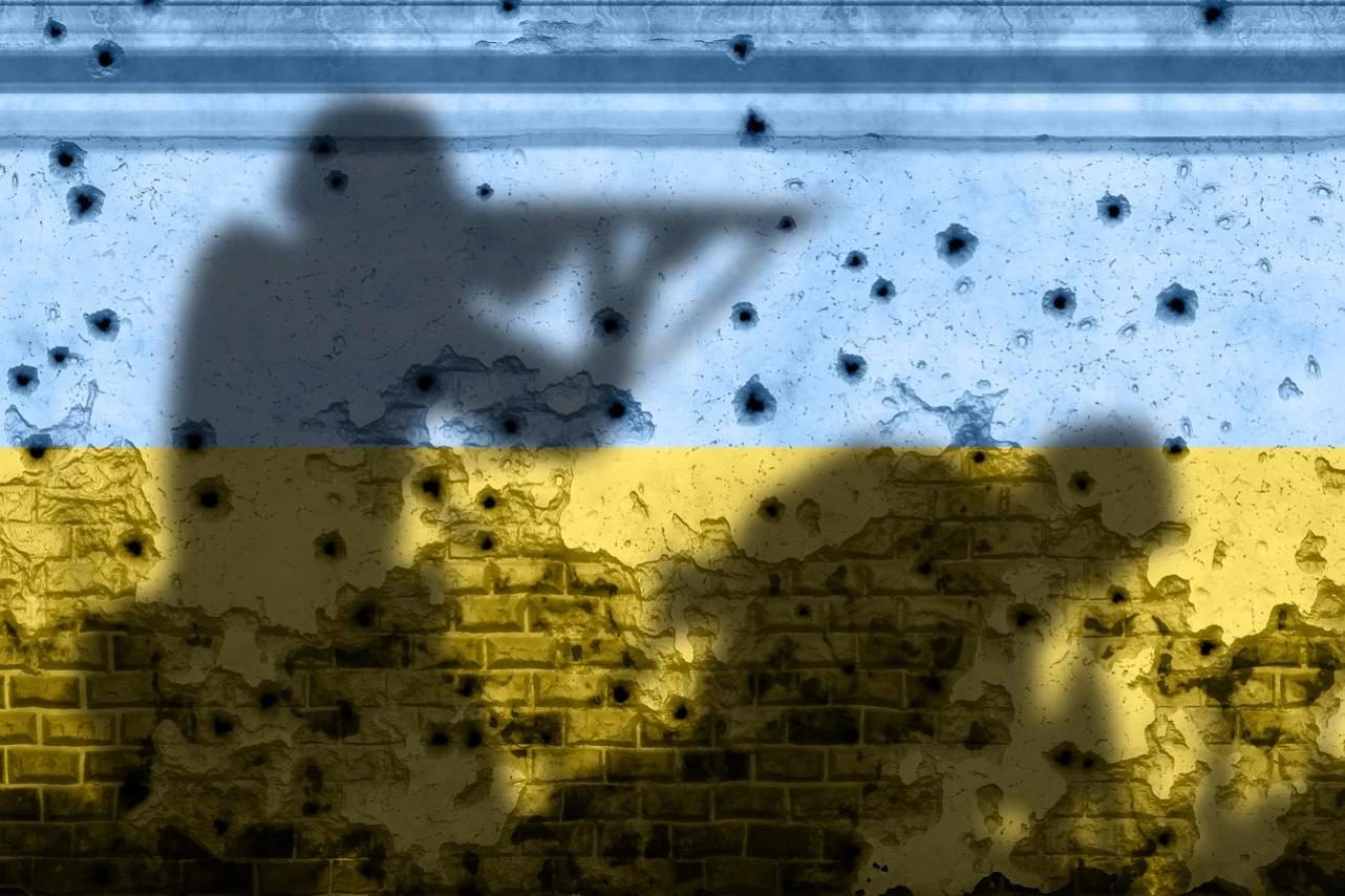 Projetées sur un grand drapeau Ukrainien (bleu et jaune) peint sur un mur, l'ombre de deux soldats. L'un des deux tient un fusil, et l'autre est courbé en avant. Le mur est parsemé d'impacts de balle.