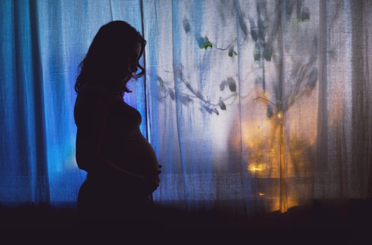 L'ombre d'une femme enceinte se détache devant un rideau fermé. L'image est assez sombre.