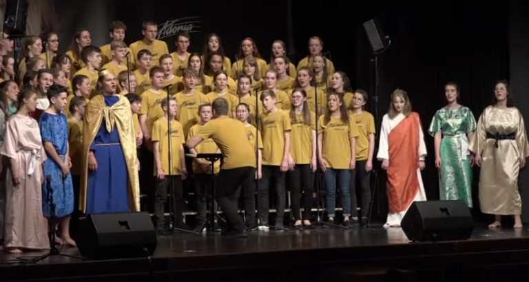 Quelques personnages en costume marchent sur une scène, devant un chœur de plusieurs dizaines de eprsonnes qui chantent. Tous sont des adolescents et des jeunes adultes.