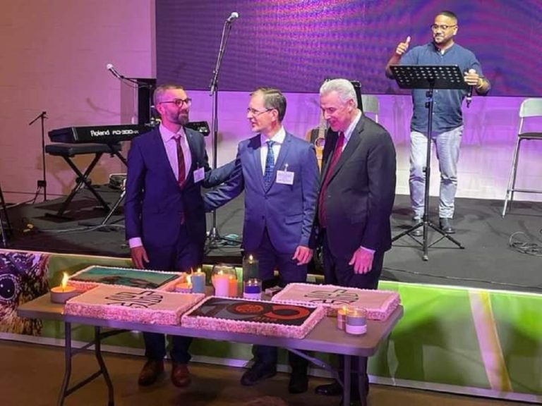 Trois délégués discutent chaleureusement devant une table où se trouvent de grands gâteau. Ils sont en intérieur, devant une scène.