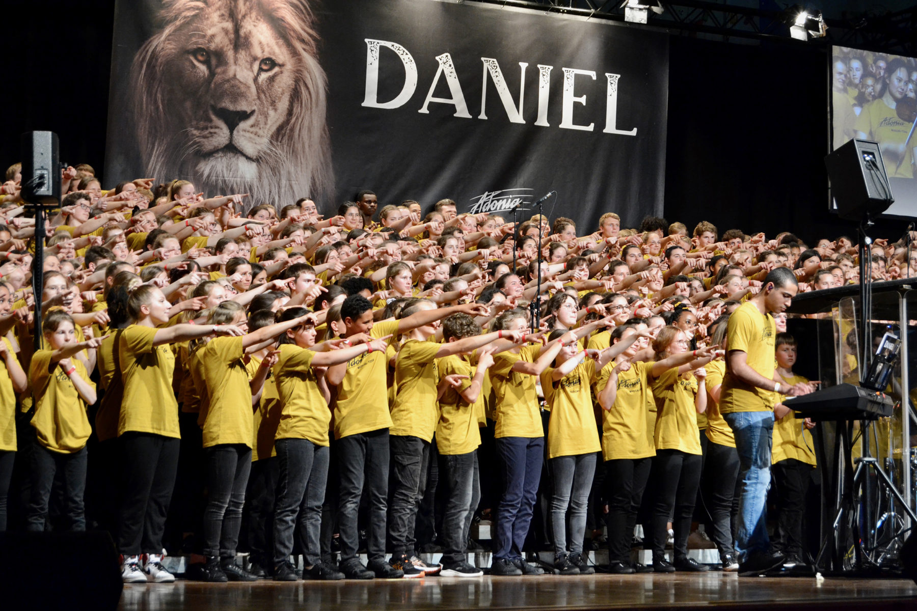 Des dizaines de jeunes, sur scène, chantent en pointant tous le public du doigt.