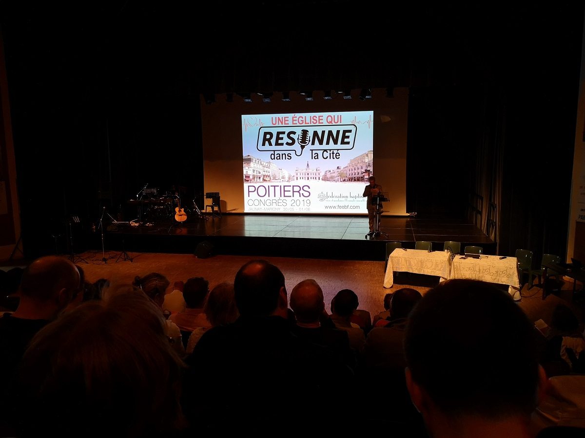 Une multitude de personnes assises dans une salle regarde vers une scène, où est projetée l'image d'introduction d'une réunion. Le slogan affiché est "Une Eglise qui résonne".