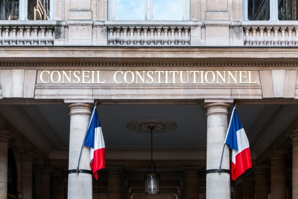 Photo de l'inscription "Conseil constitutionnel" sur l'entrée du bâtiment qui l'abrite. Deux drapeaux français sont dressés de chaque côté, sur des colonnes.