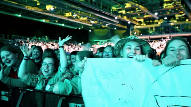 Une foule de jeunes dans une salle de concert. Au premier plan, des jeunes filles crient, rient et tiennent une banderole.