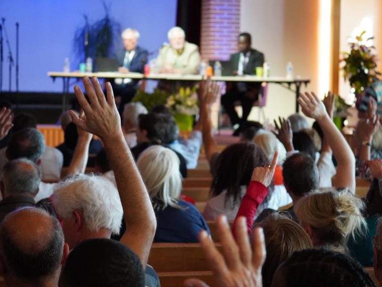 Des hommes et des femmes lèvent la main pour donner leur accord lors d'une Assemblée générale. trois personnes sont assises à une table sur une scène.