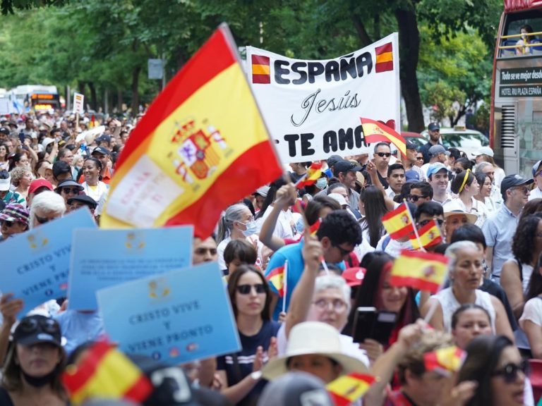 Des femmes et des hommes tiennent des drapeaux espagnols et des pancartes où sont écrit le nom de Jésus. Ils sont en train de marcher dans une large avenue de Madrid.