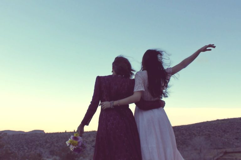 Deux femmes en robes se tiennent joyeusement par la taille, en campagne, à l'aube. L'une d'elle tient un bouquet de fleurs.