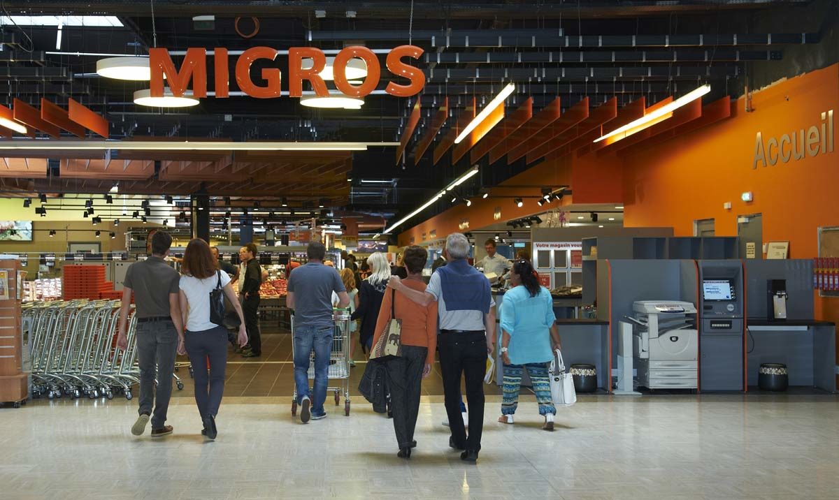 Plusieurs personnes entrent dans un magasin Migros avec des caddies.