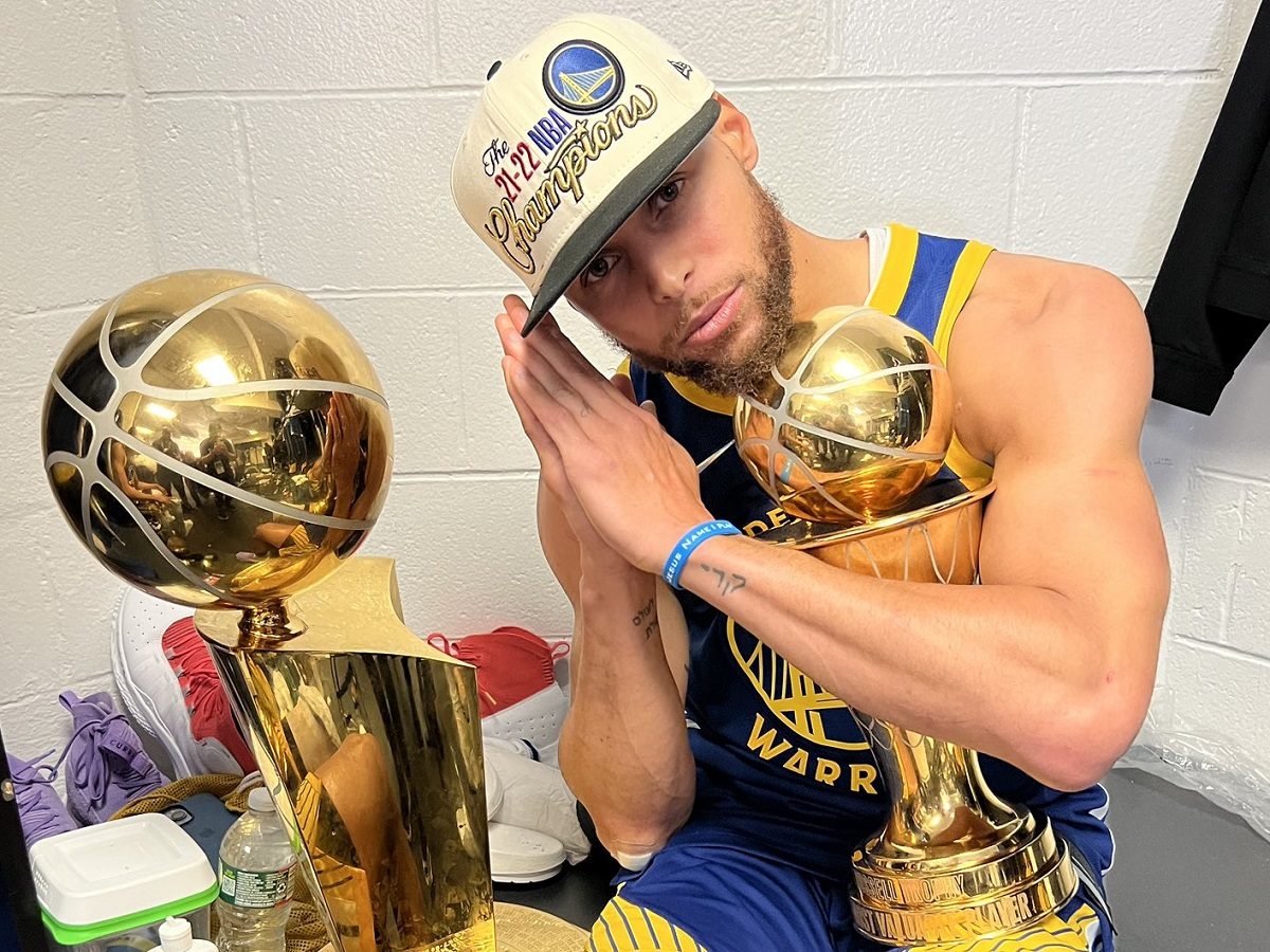 Le basketteur Stephen Curry avec la casquette de champion NBA, le trophée doré et le titre MVP des finales. Il fait un signe de prière.