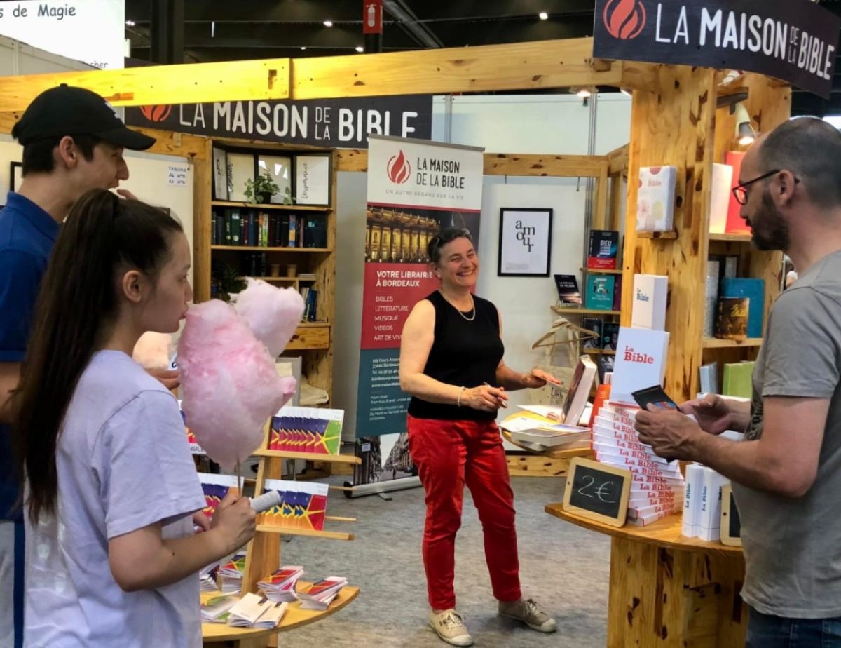 Une bénévole anime le stand de la Maison de la Bible, entourée de livres. Plusieurs personnes écoutent.