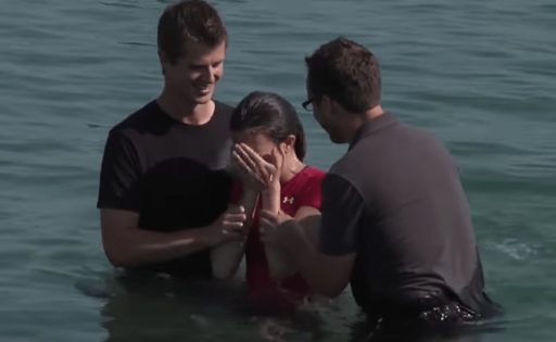 Deux personnes baptisent une femme en extérieur. Ils ont de l'eau jusqu'à la taille et semblent joyeux.
