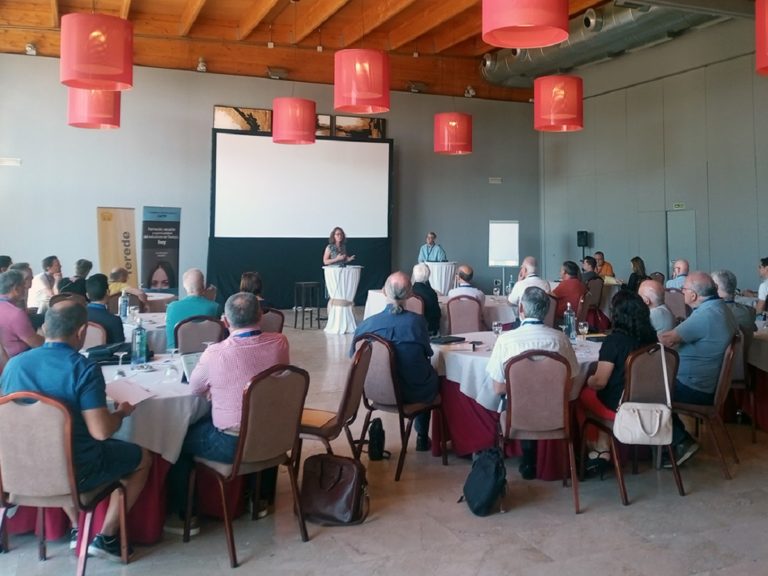 Une trentaine de personnes assises à plusieurs tables dans une salle écoutent deux orateurs.