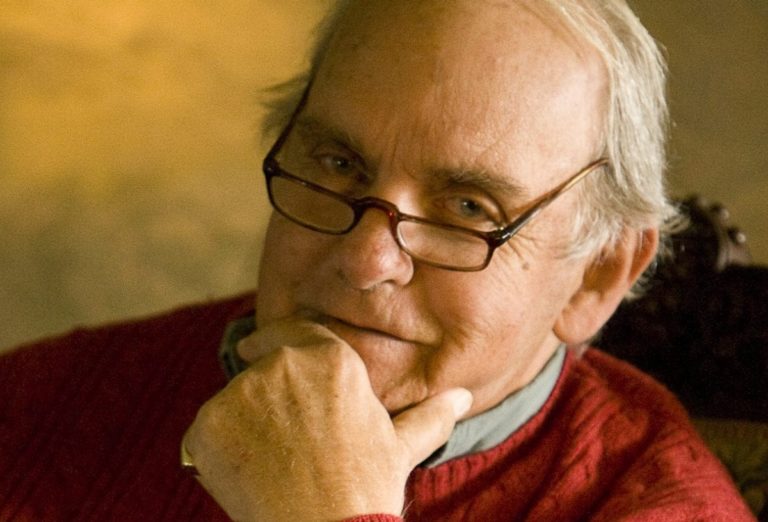 L'écrivain Frederick Buechner, en pull rouge, main sur le menton