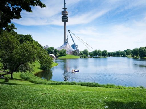 De la pelouse et des arbres autour d'un lac avec en arrière plan le parc olympique de Munich et sa grande tour