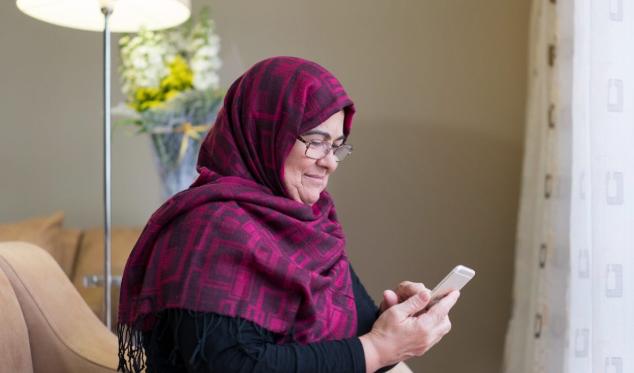 Une femme un peu âgée portant un hidjab regarde son smartphone en souriant. Elle est seule, assise sur un canapé.
