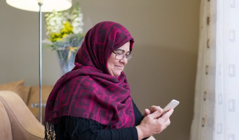 Une femme un peu âgée portant un hidjab regarde son smartphone en souriant. Elle est seule, assise sur un canapé.