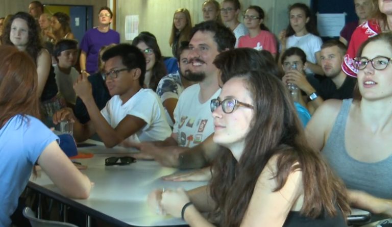 Des étudiants joyeux sont assis à des tables de cantine. Ils écoutent quelqu'un parler.