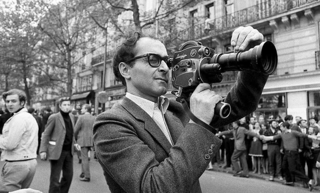 Jean-Luc Godard filme dans une rue. Les passants sont retenus sur les trottoirs.