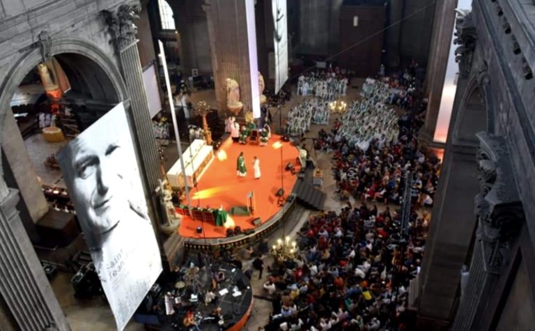 Une immense foule est assise dans une vaste église, à l'écoute d'un prêtre.