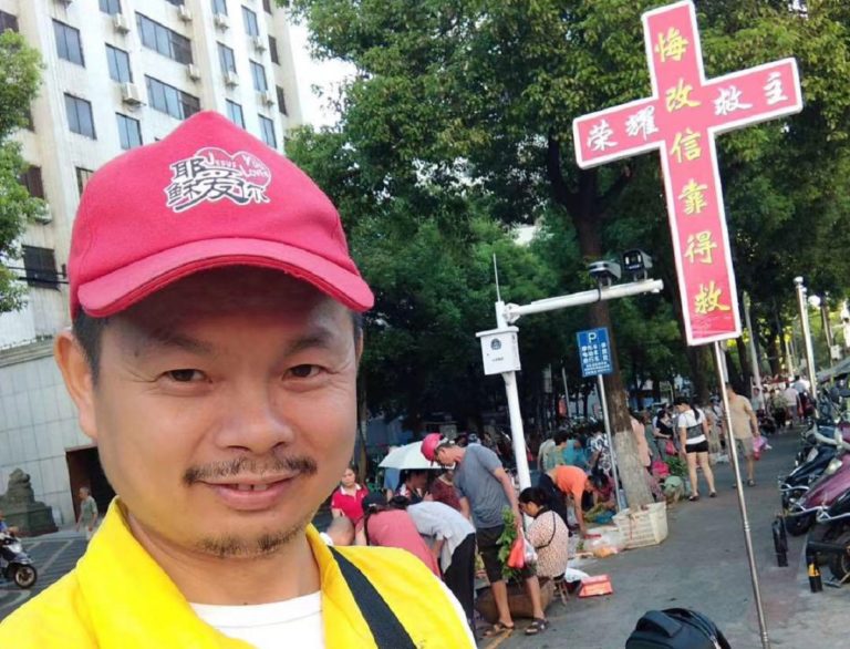 Chen Wensheng avec une casquette rouge et l'inscription "Jésus est Amour" tient un stand d'évangélisation avec une grand croix rouge et jaune