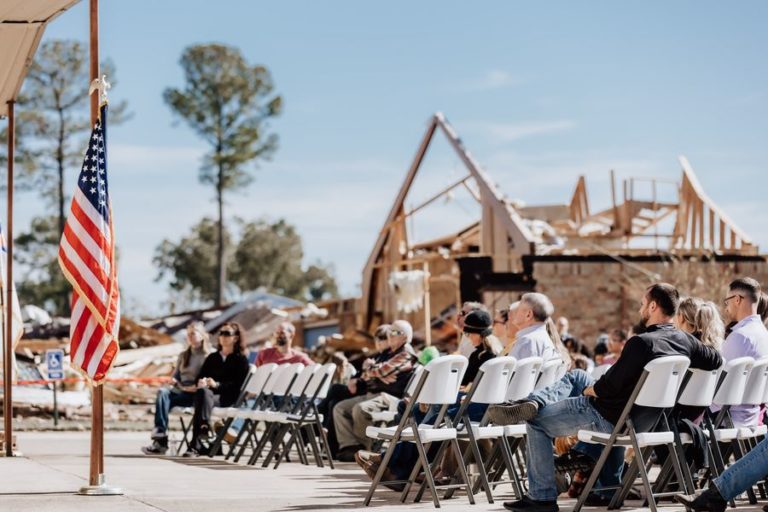 des chrétiens assis sur des chaises louent Dieu en plein air après la destruction de leur Eglise en arrière plan