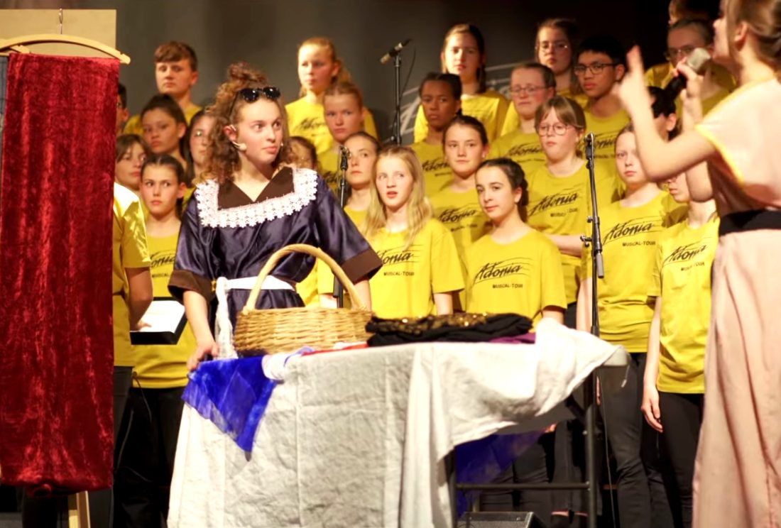 Deux jeunes comédiennes jouent au premier plan durant un spectacle. Derrière elle, une chorale suit la scène.