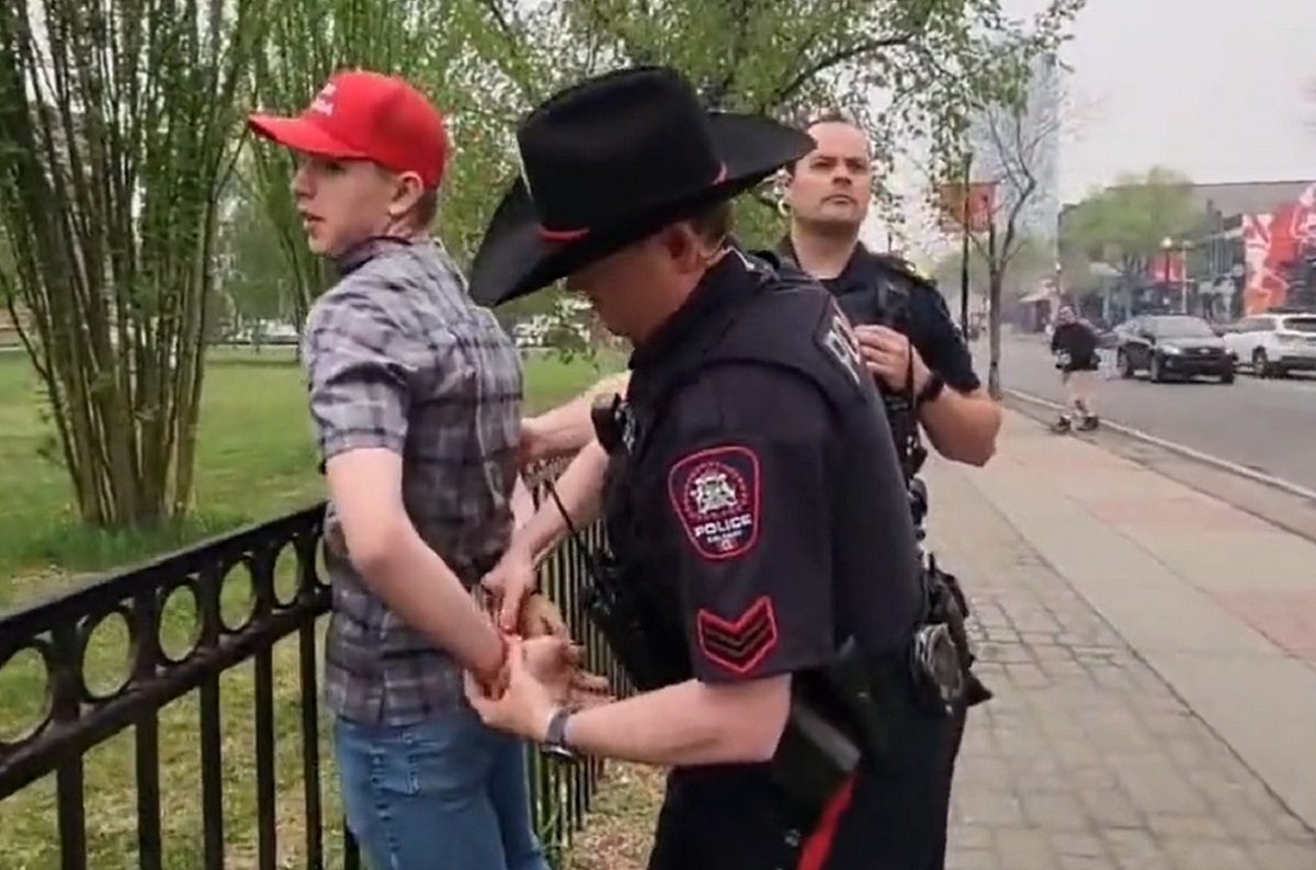 Portant une casquette rouge Josh Alexander, les mains dans le dos, est menotté par un policier avec un chapeau noir