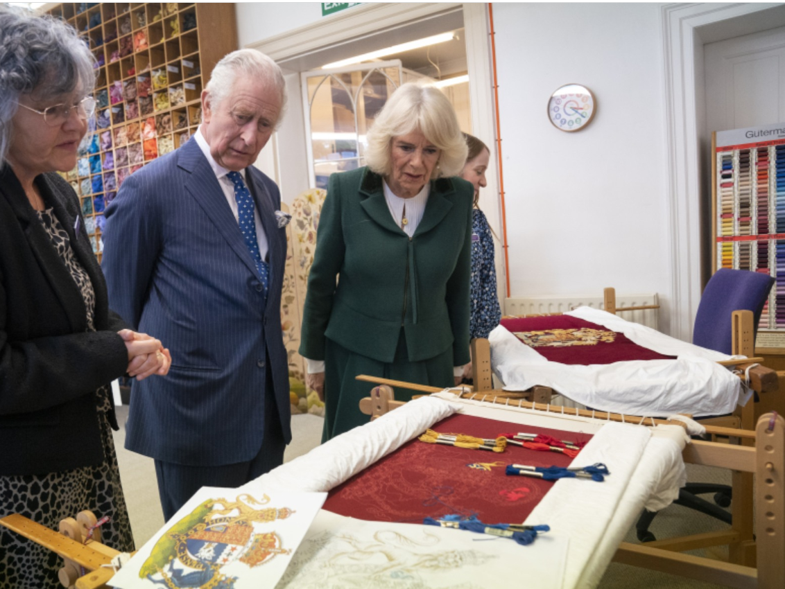 Charles III et son épouse Camilla regardent des tissus et broderie étalées devant eux.