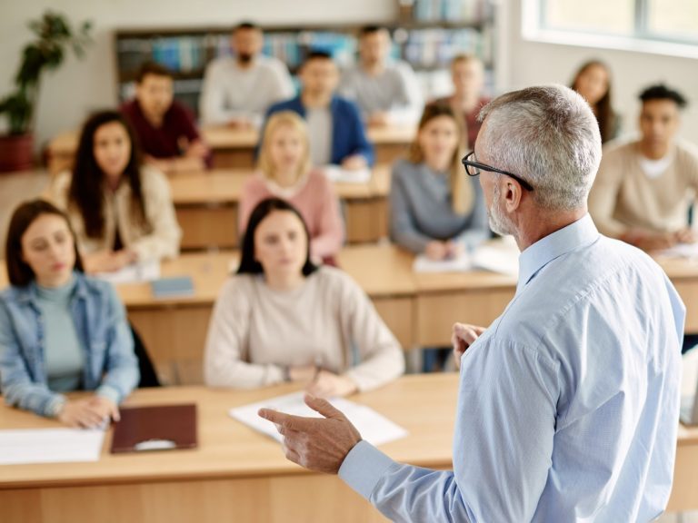 Un professeur aux cheveux gris donne un cours à des élèves assis derrière leurs bureaux en bois