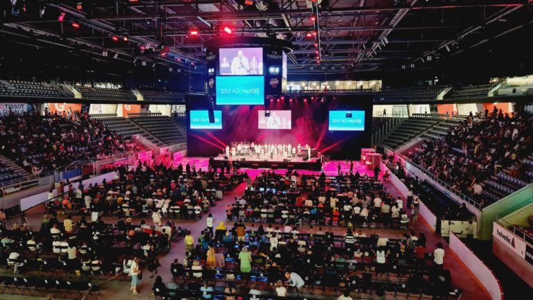 Des centaines de personnes assises dans une immense salle regardent vers la scène, où se tient un groupe d'intervenants.