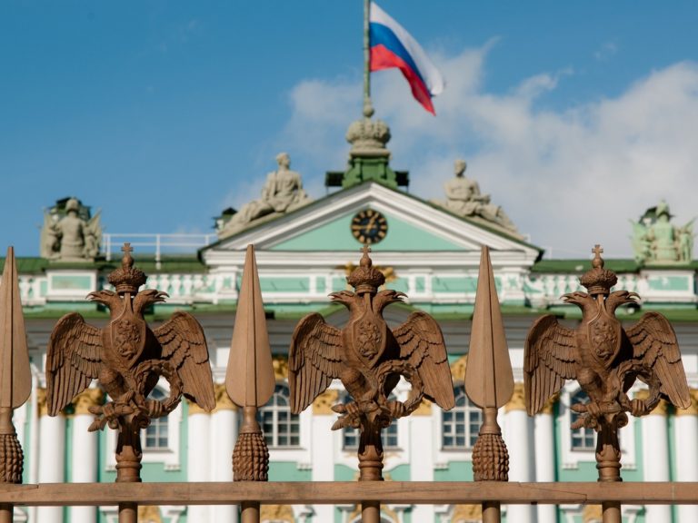 Au premier plan, une grille l'aigle bicéphale russe ; au second plan un bâtiment avec un drapeau russe à son sommet
