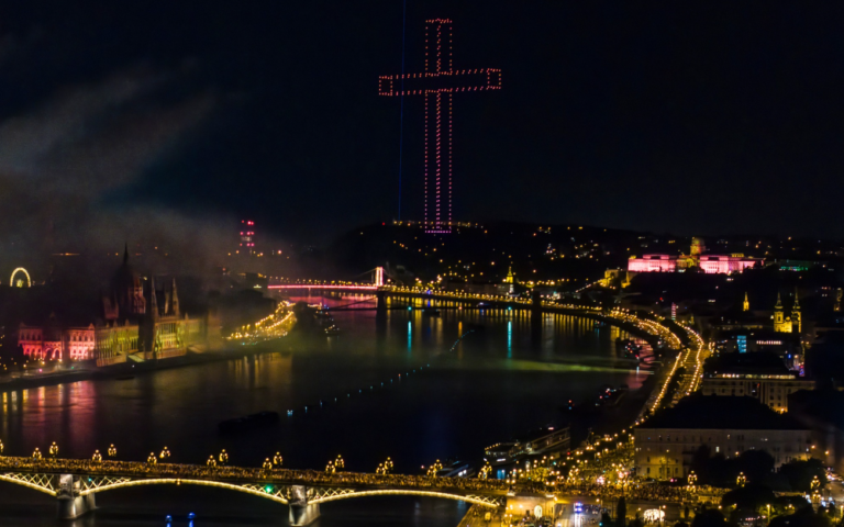 Une immense croix dans le ciel formée de points lumineux. En dessous, la ville de Budapest illuminée dans la nuit.