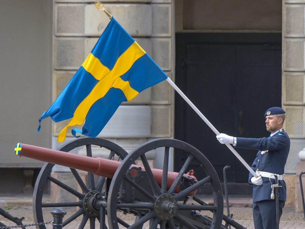 Un homme tient un drapeau suédois, bleu à croix jaune, près d'un canon